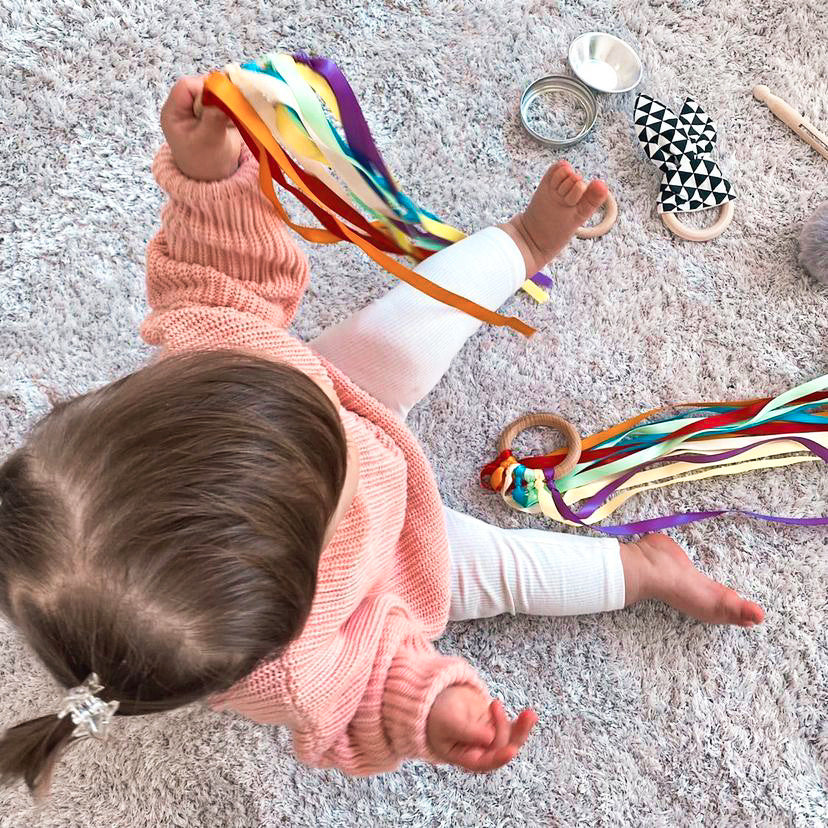 baby sitting on the floor playing with a rainbow ribbon toy and heuristic play items